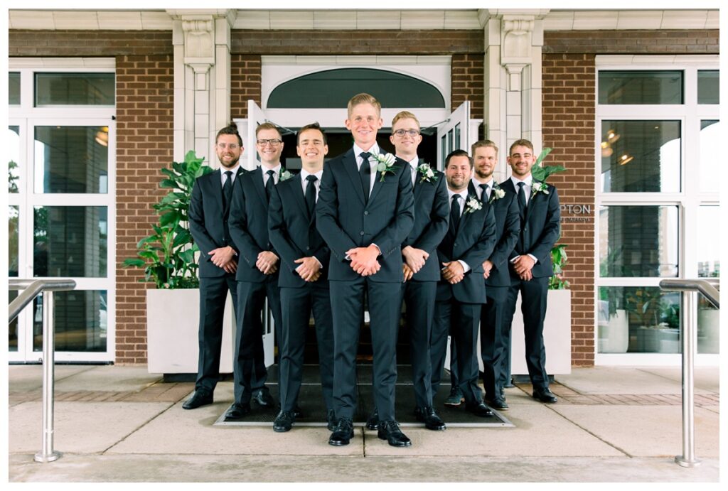 Group photo of groom and groomsmen smiling in front of the hotel
