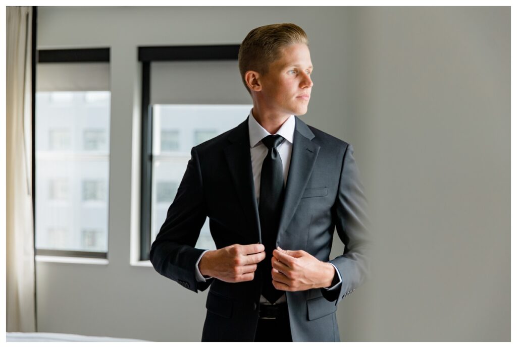 Groom buttoning his suit jacket by window