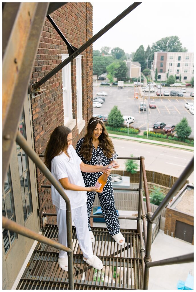 Bride opening champagne off fire escape with her sister