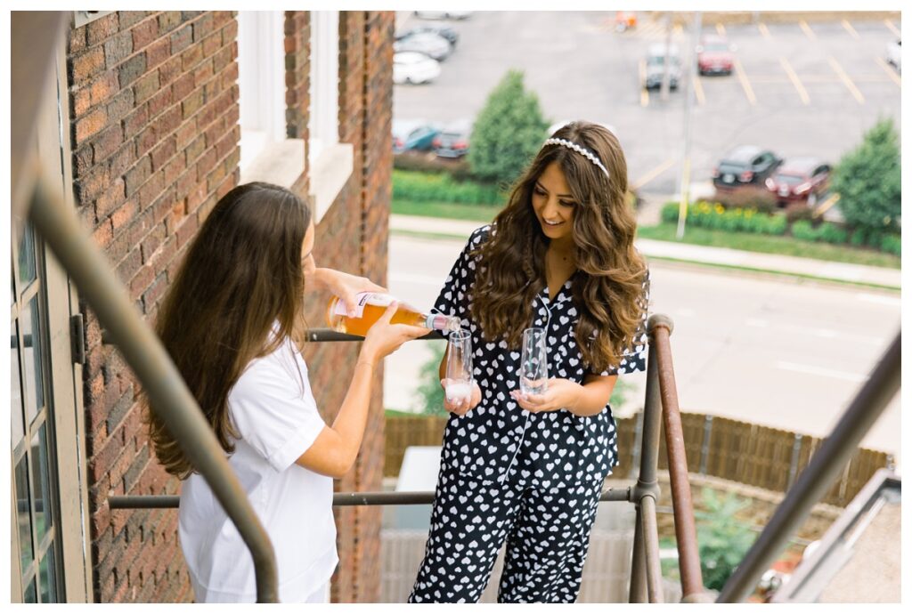 Bride pouring champagne into flute with sister on hotel fire escape
